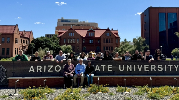 A group of people posing around the Arizona State University sign on the ASU Tempe campus with Old Main building in the background