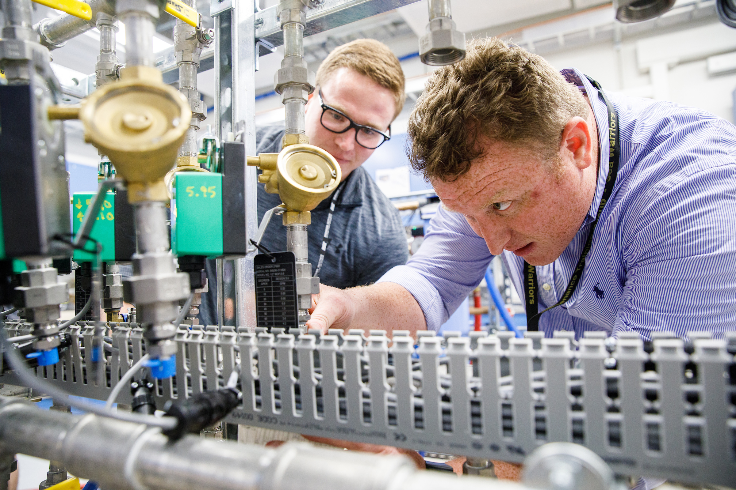 Two young men peer at an array of scientific components. The caption: Students Alex Gardeck and Brandon Cook examine the precision thermal trim unit water systems that controls the temperature of components in the radiography/fluoroscopy room. 