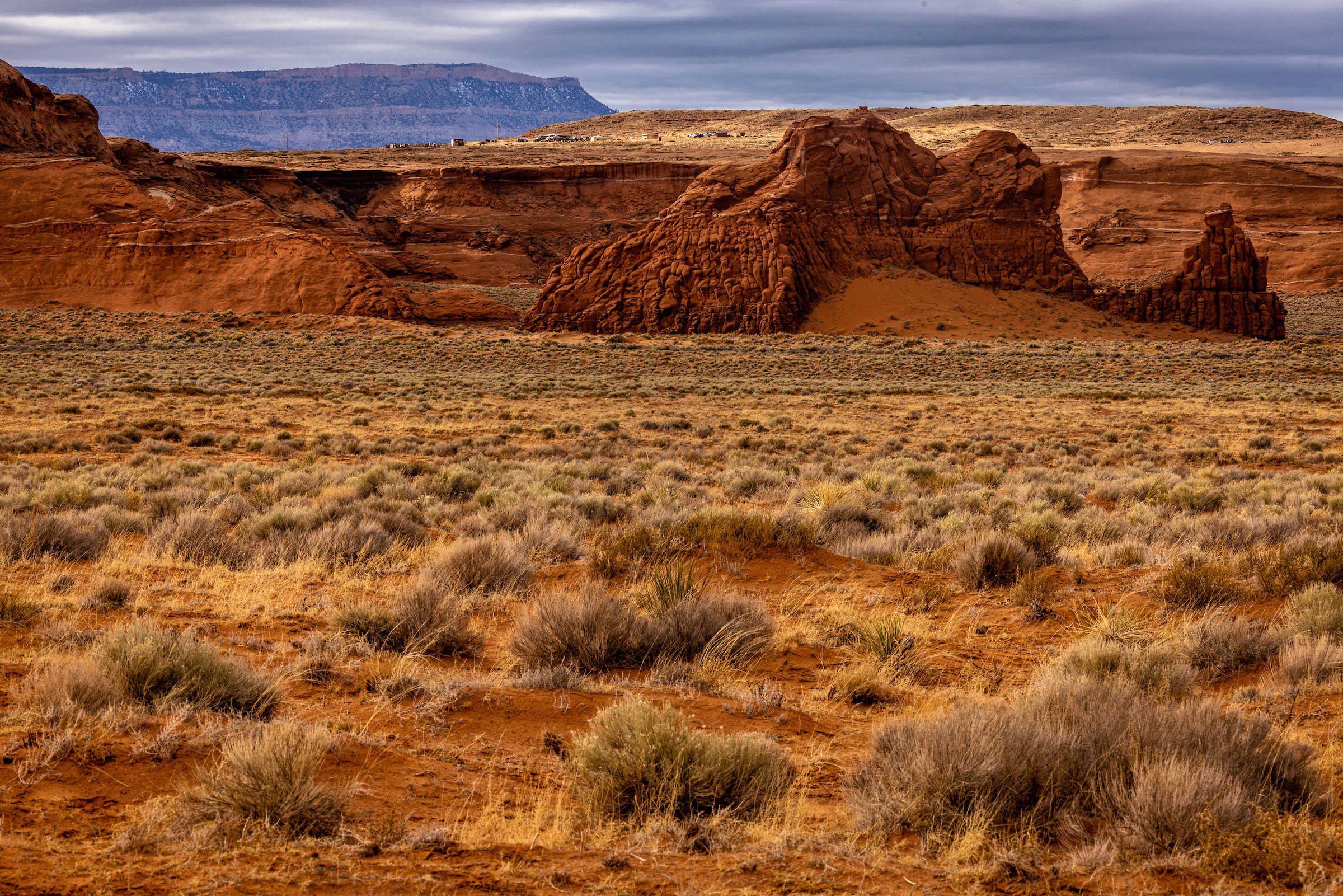 Structures and vehicles can barely be seen in the distance on a mesa in northern Arizona with sculpted red hills in the foreground