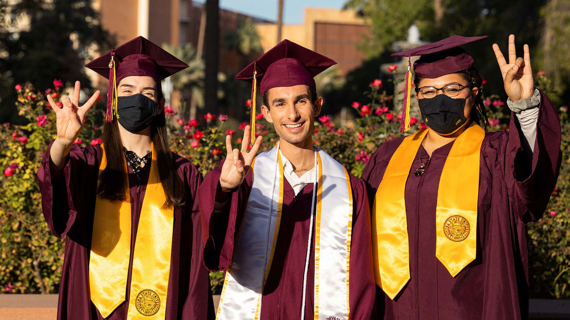 Three students in graduation gowns and caps make the pitchfork gesture; one is unmasked and two have masks on