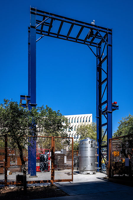A blue gantry stands high above a big metal canister and other mechanical equipment outside