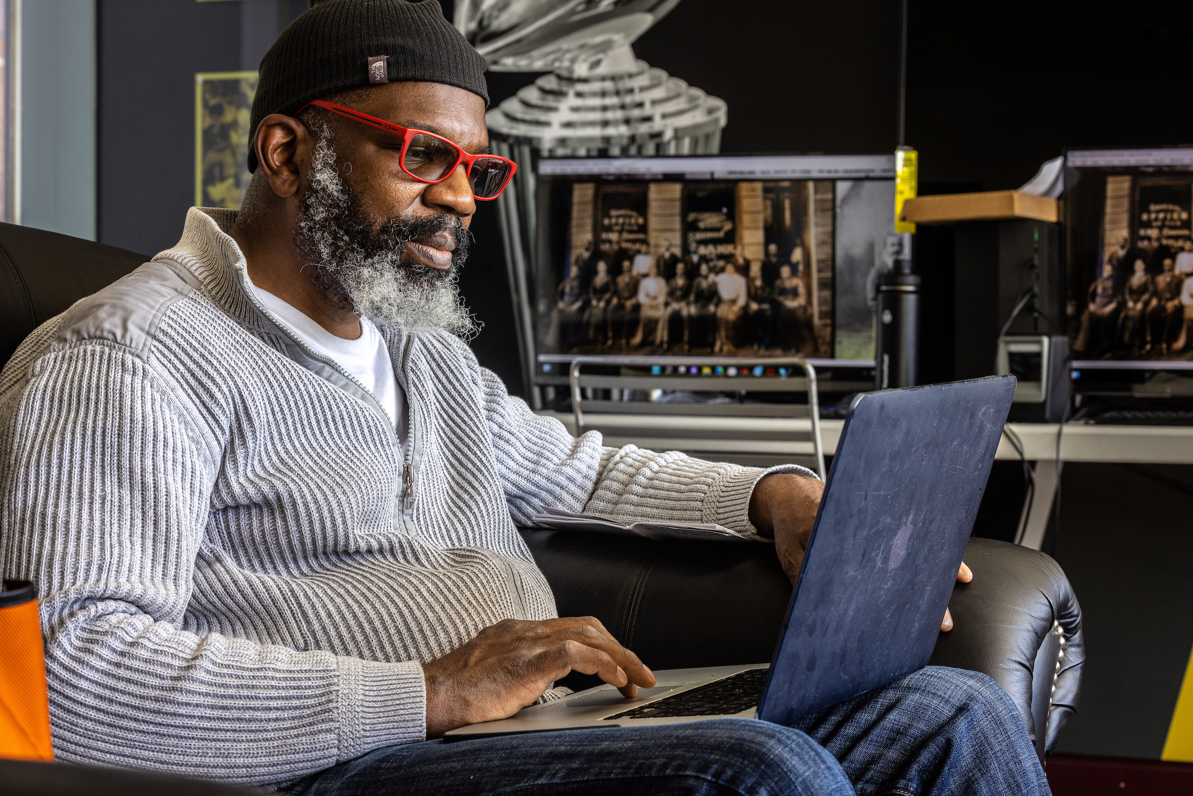 Black man wearing hat and red glasses sitting in chair looking at laptop
