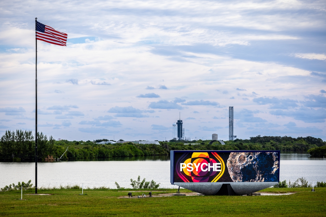 A sign with Psyche imagery in the foreground with NASA launch sites in the background