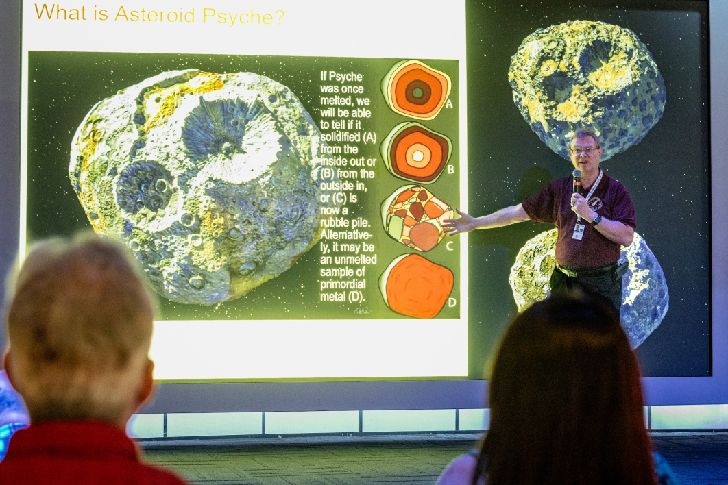 A man speaks to a crowd in front of a display showing asteroids