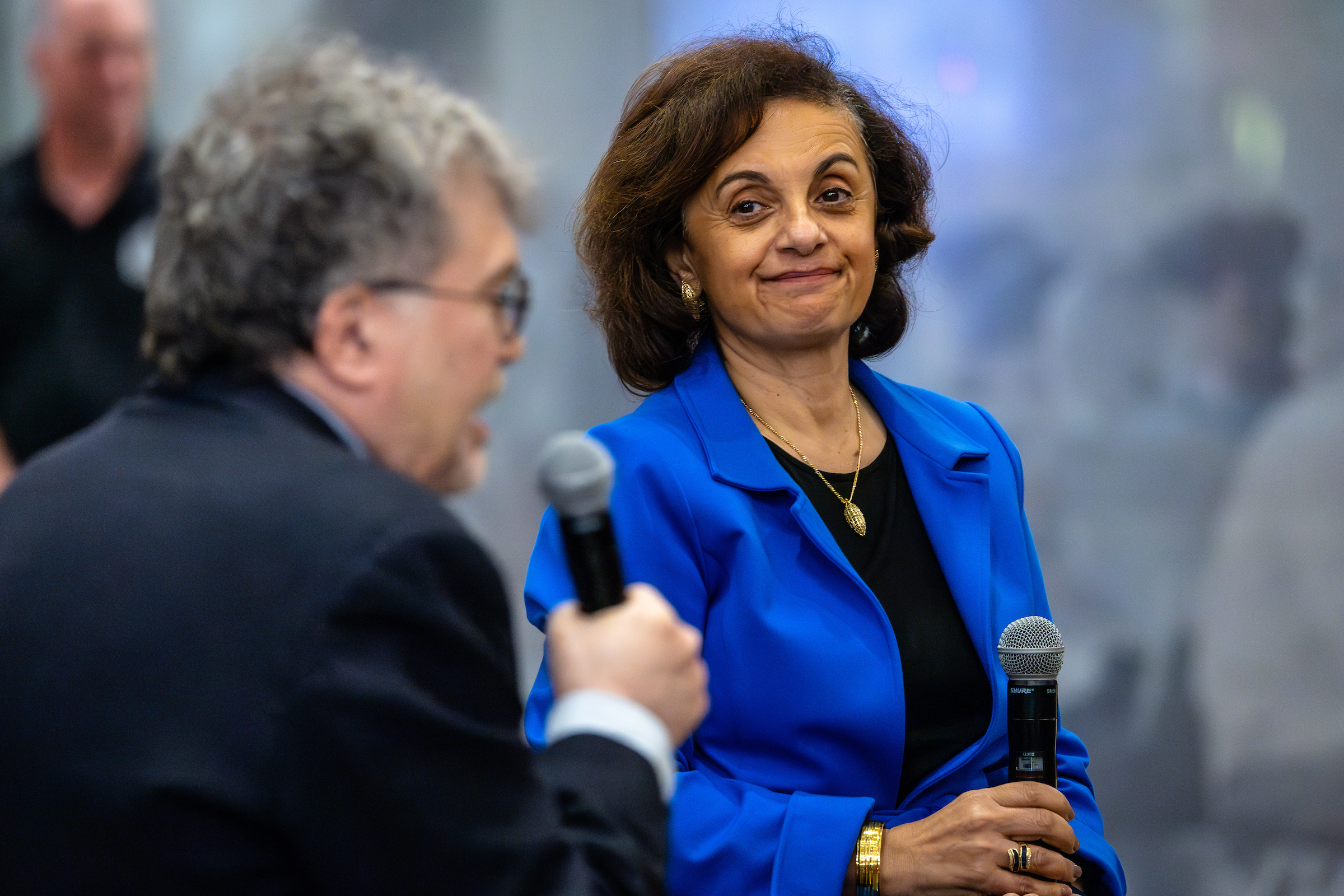 Woman with short brown hair and blue jacket holds microphone and listens to man holding microphone speak