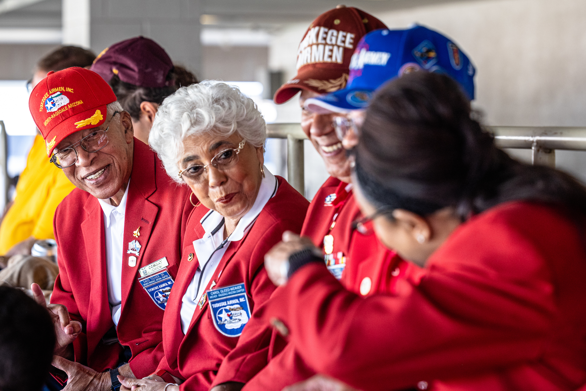 People in Tuskegee Airmen hats and red blazers chat in stadium seats