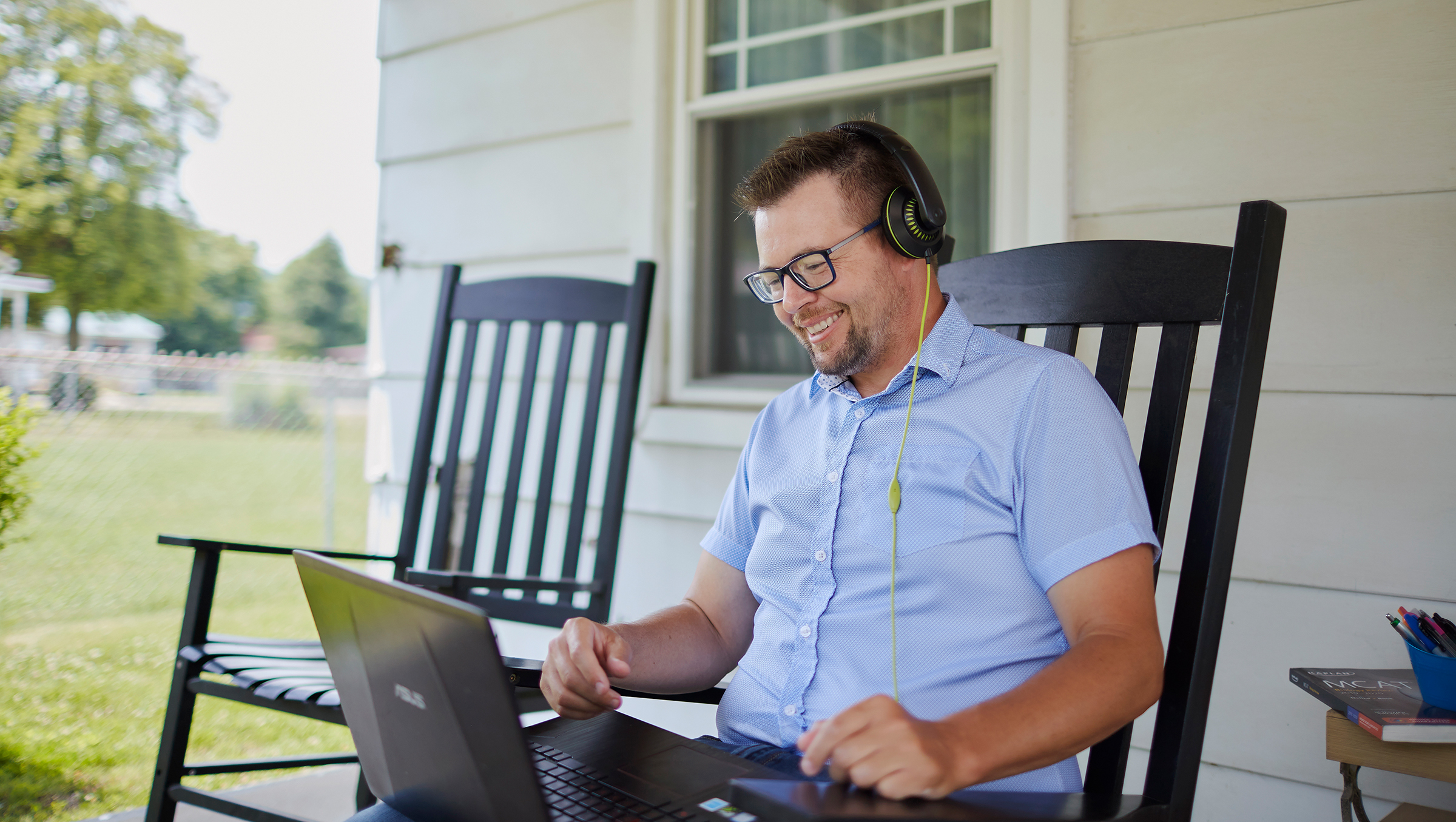 A man sitting in a rocking chair on a porch works on a laptop while wearing headphones