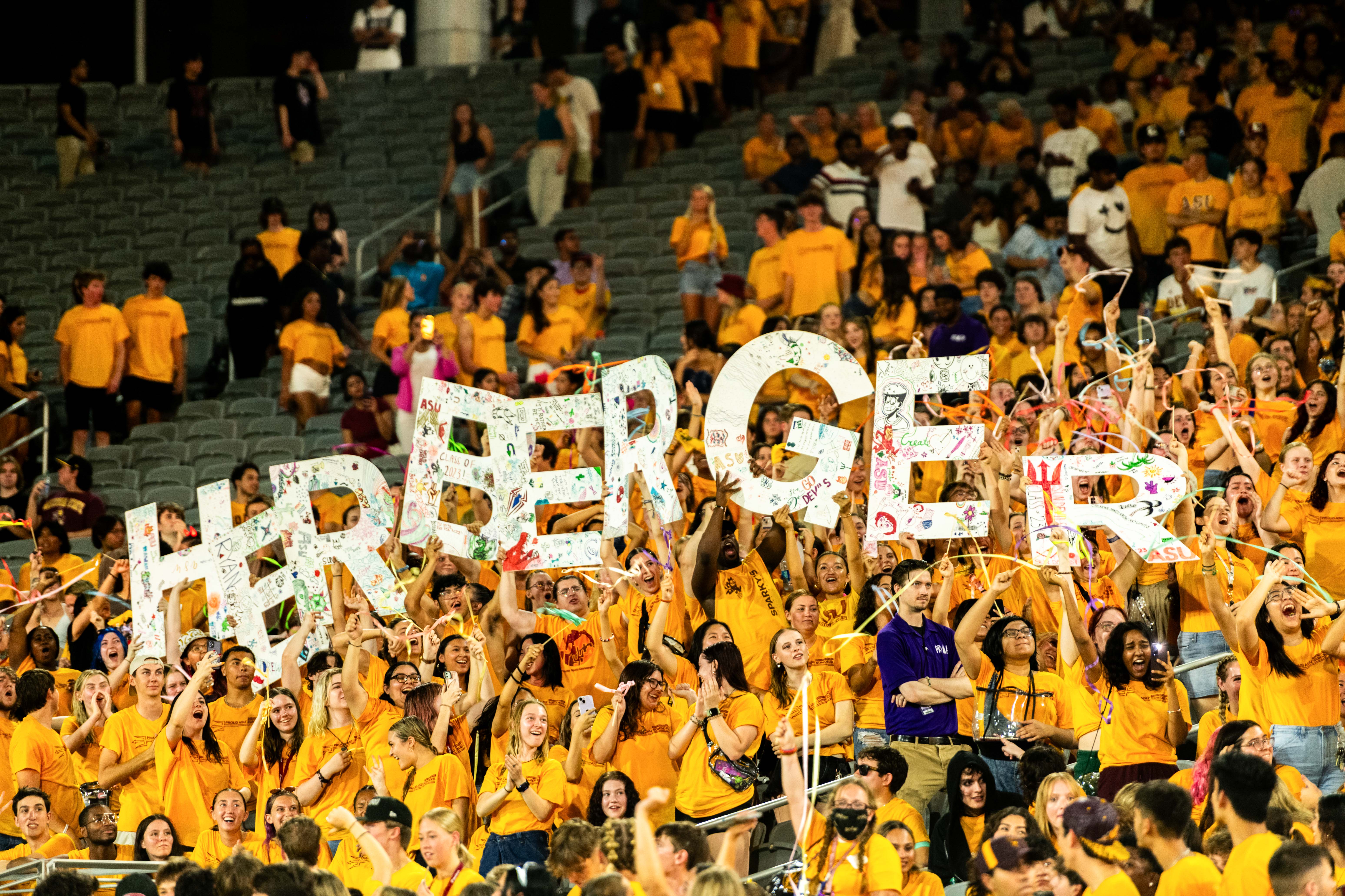 Group of students in football stadium wearing gold t-shirts and holding up letters that spell Herberger