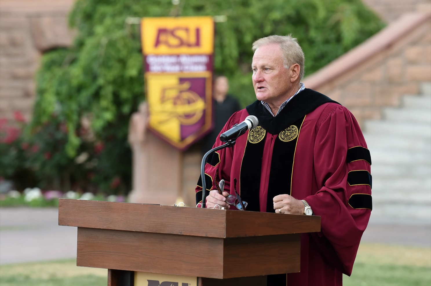 Man wearing graduation regalia standing behind a lectern and speaking into a microphone.