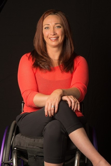 Portrait of a woman with long brown hair wearing a coral shirt and black pants, seated in a wheelchair and smiling at the camera.