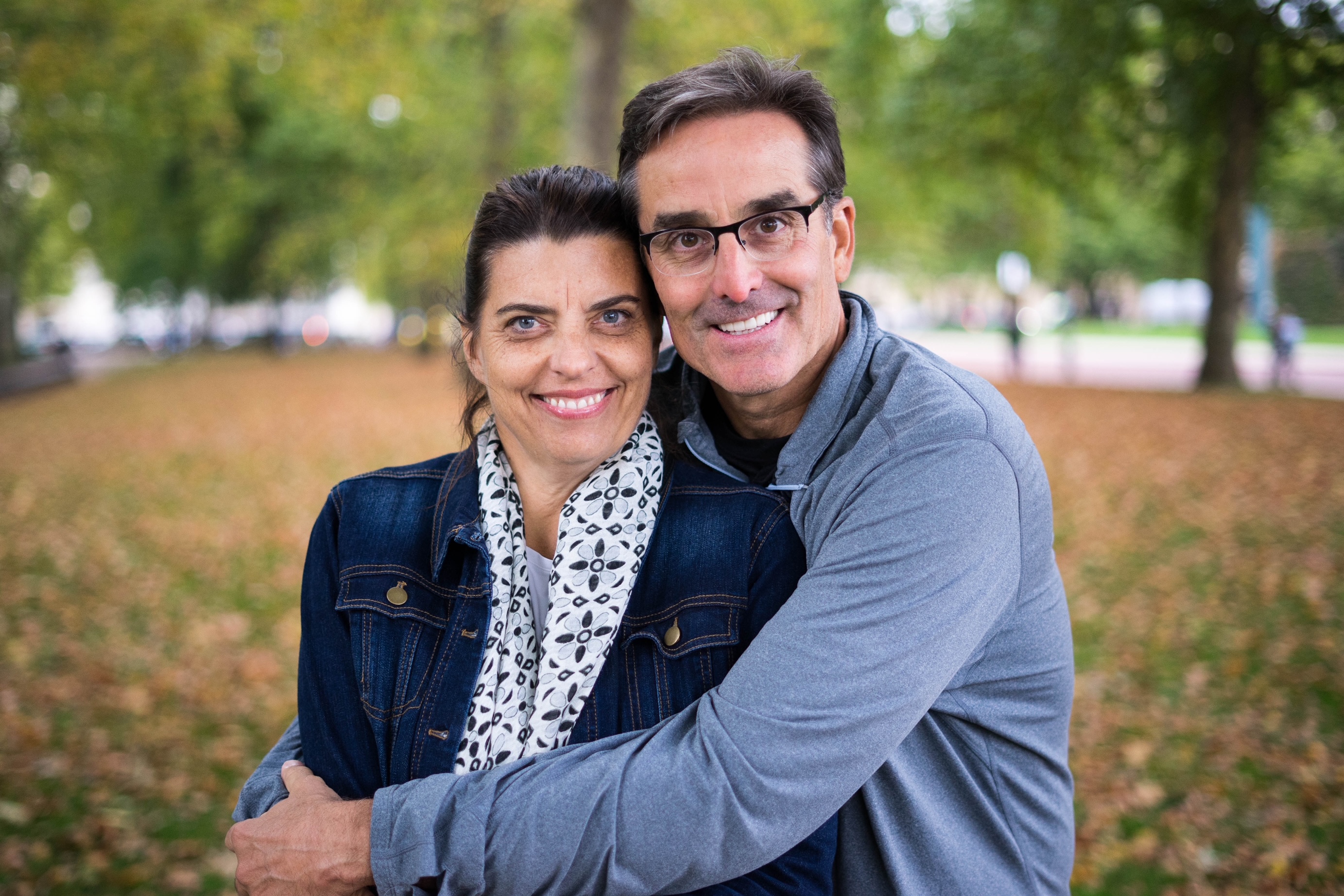 a woman and a man embrace and smile at the camera with a fall foliage scene in the background