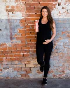 A woman stands against a brick wall, holding a bottle of juice