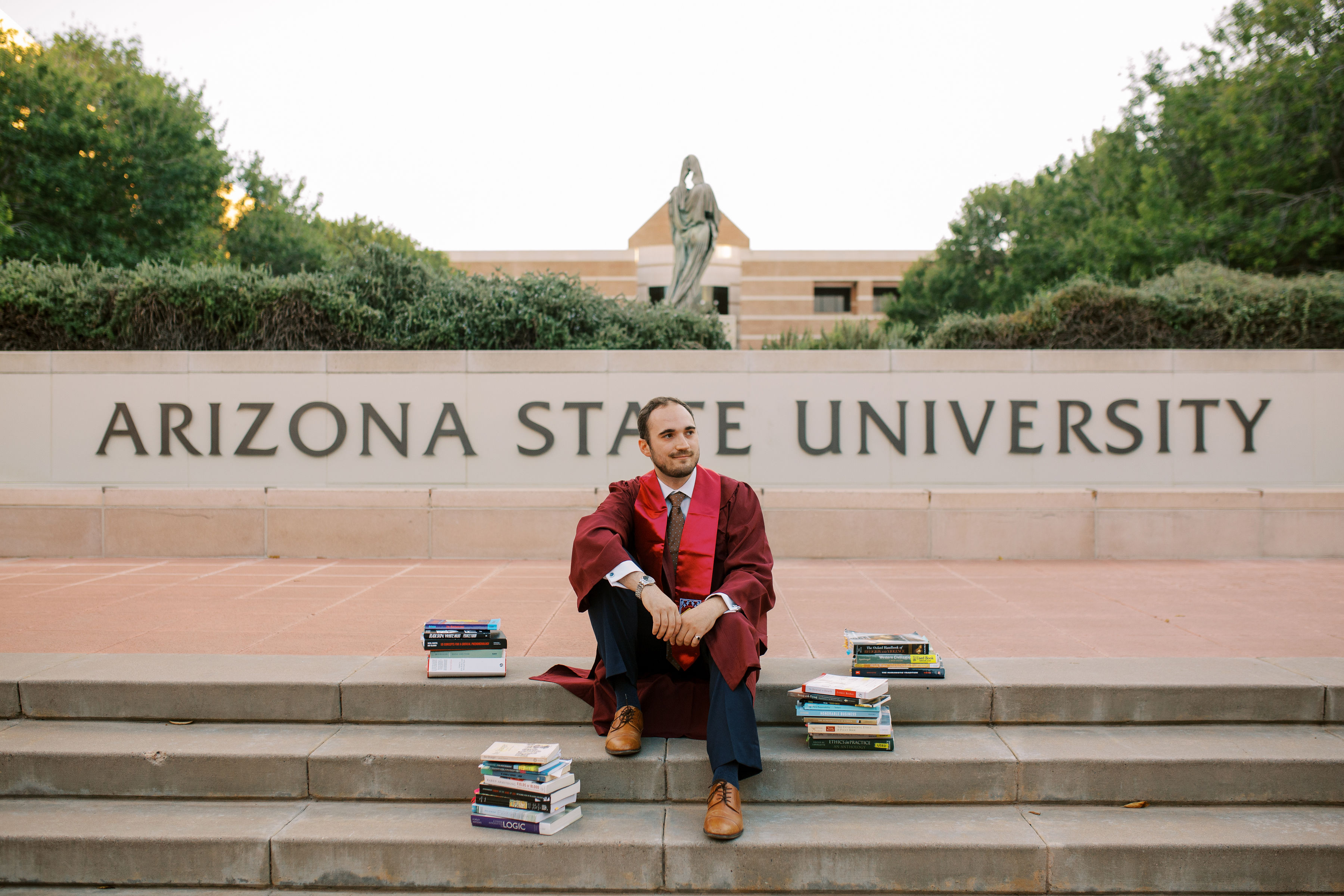  sitting on steps in front of Arizona State University in graduation gown surrounded by books