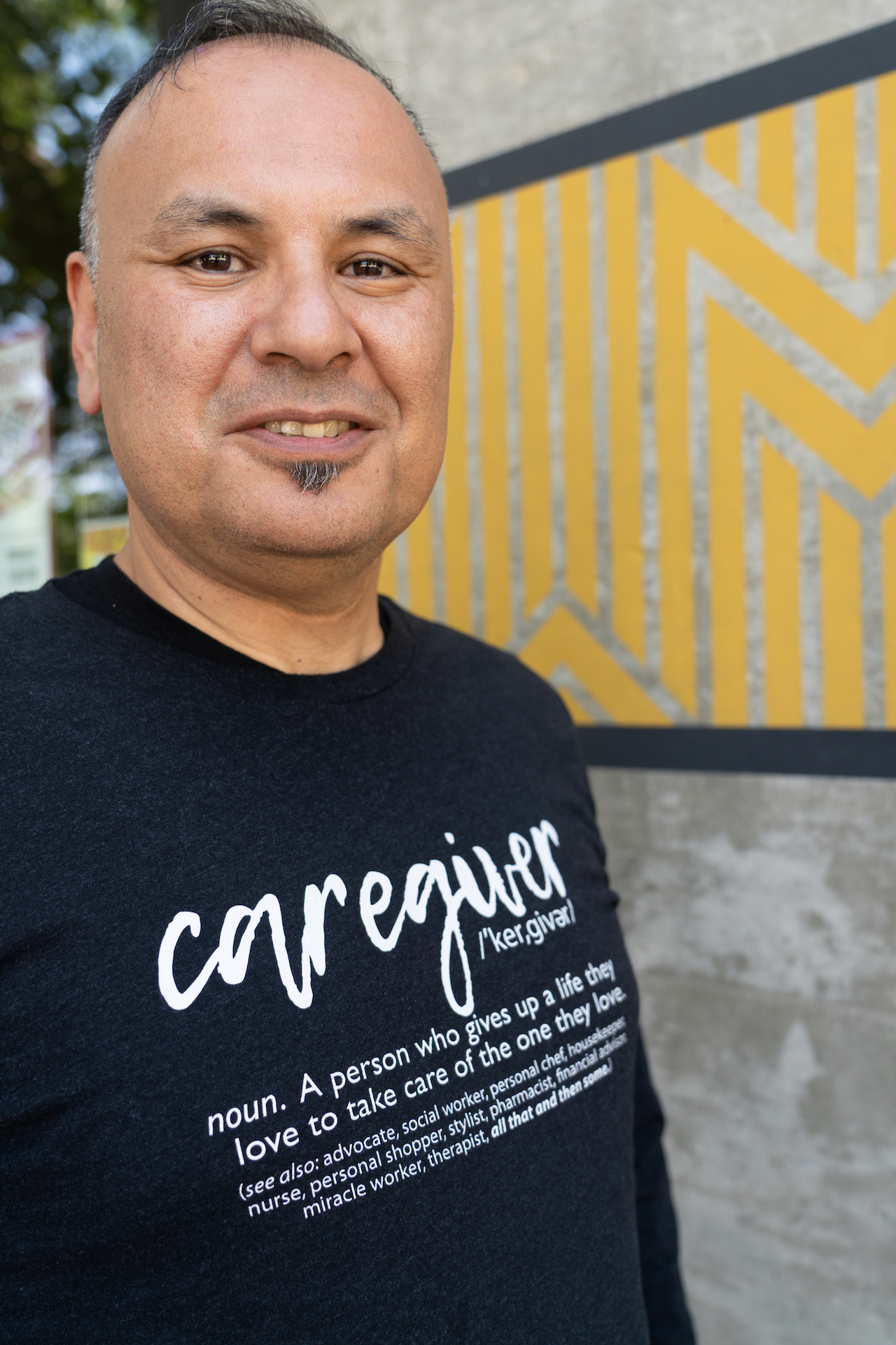 Carlos Olivas looks at the camera. He's wearing a black shirt with the word caregiver on it and the definition below.