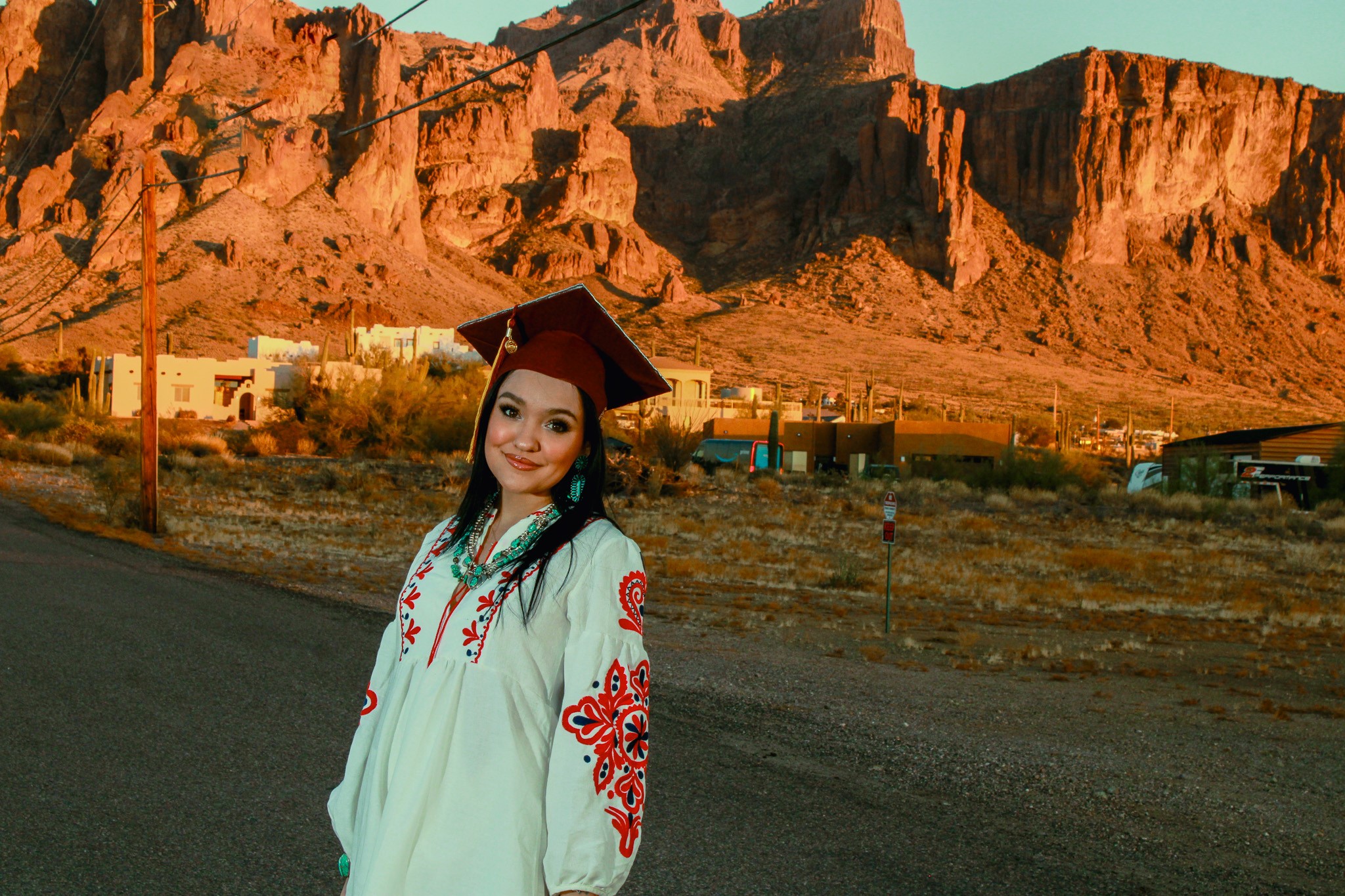  smiles at the camera wearing her ASU graduation cap and a white shirt with red flowers on the sleeves. A mountain range can be seen in the background