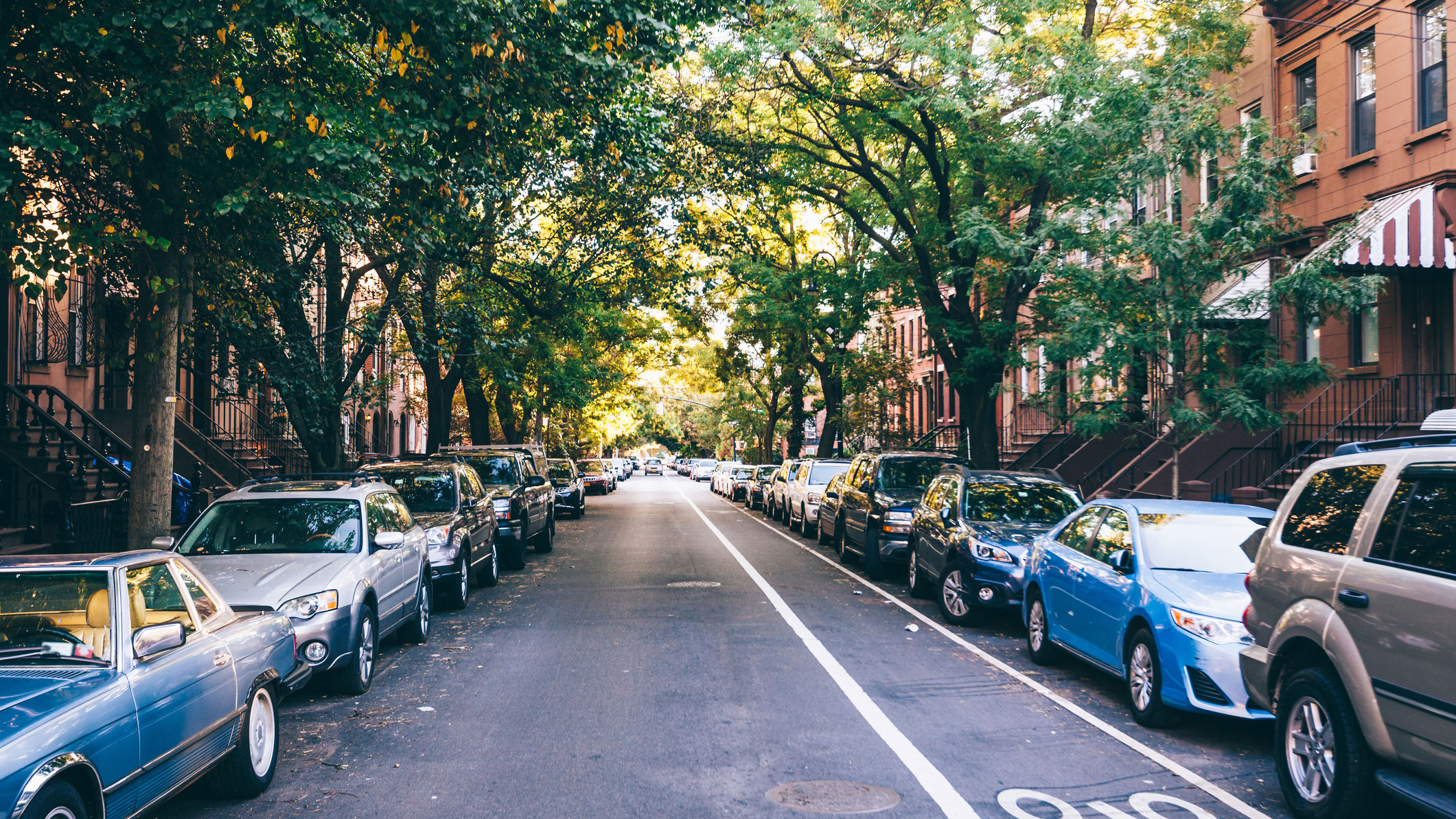 A street in a neighborhood of brownstones with the road lined with cars on both sides