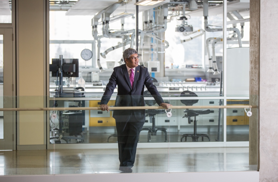 University of Calgary Professor Janaka Ruwanpura wearing a suit and leaning against a railing in front of a window overlooking a lab space.