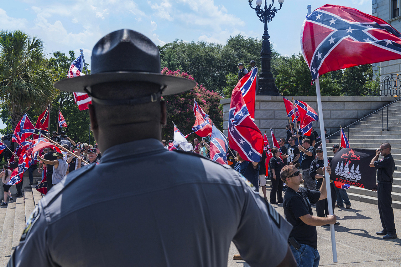 People carrying confederate flags and KKK banners protest at a government building as a Black guard looks on