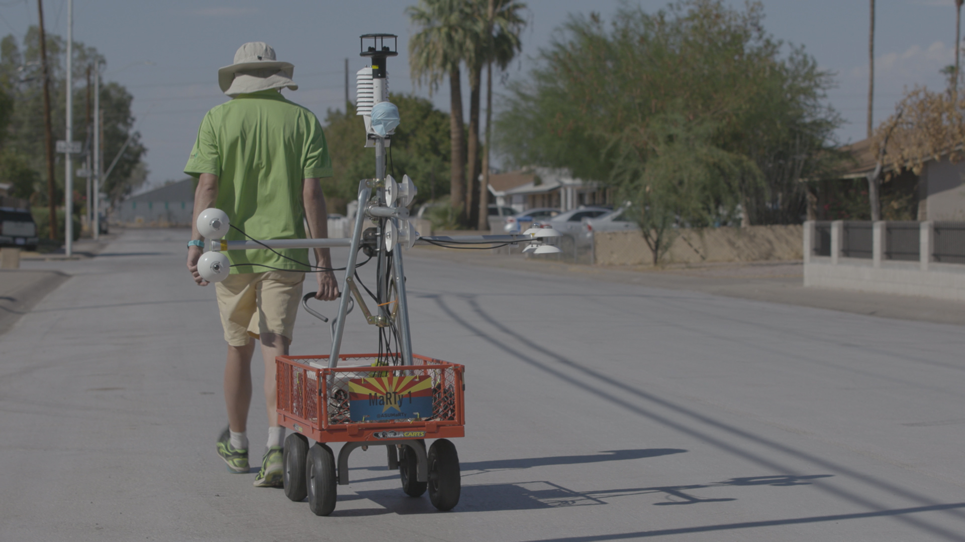 A man wearing a sun hat pulls a cart with a bunch of temperature sensors down a neighborhood street