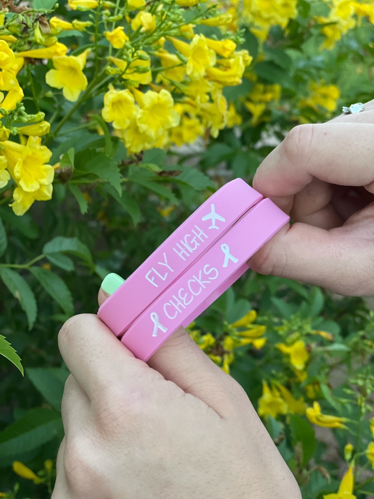 Hands hold two pink wristbands with the words Fly High and Checks on them along with breast cancer ribbon icons