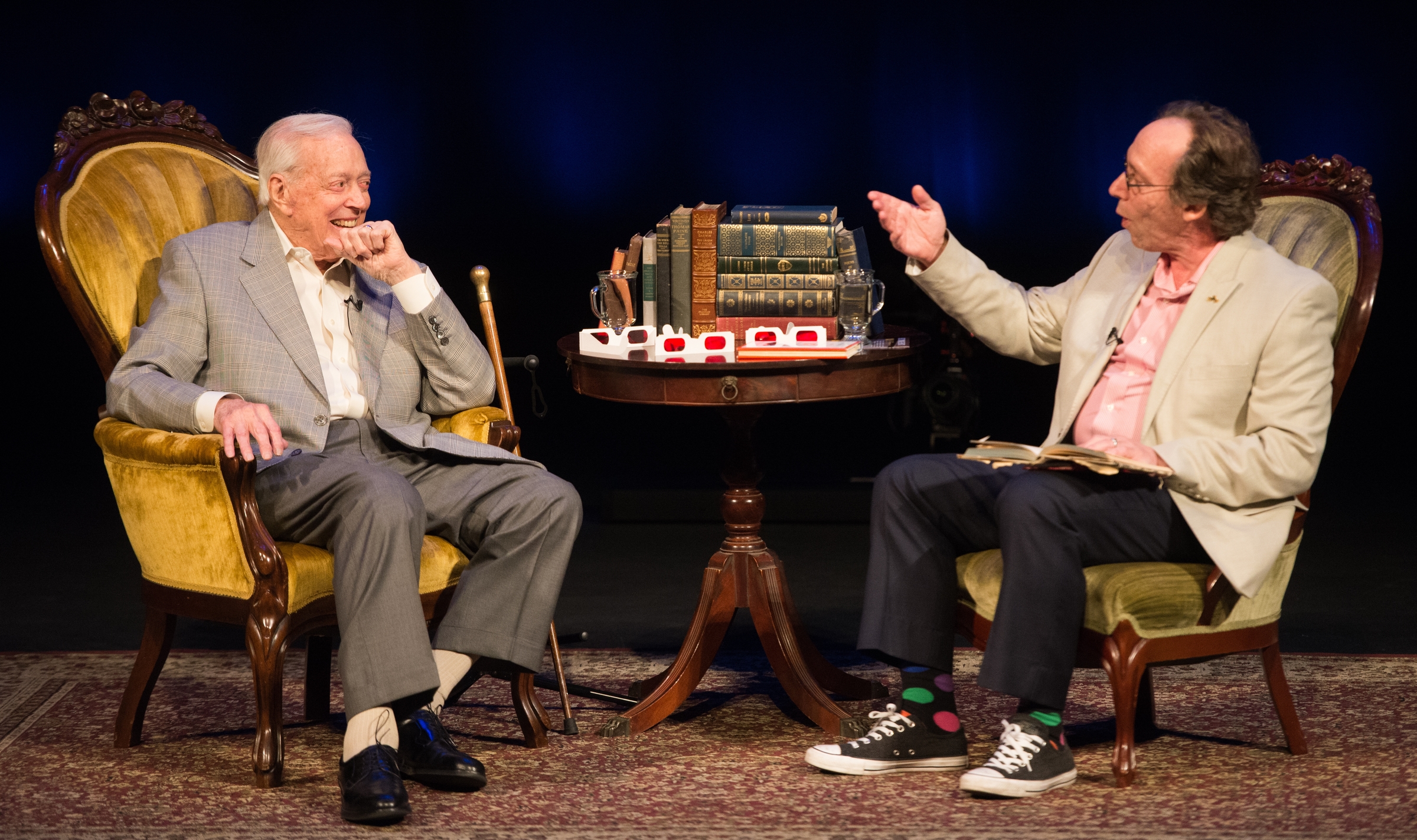 Two men sit in armchairs on a stage.