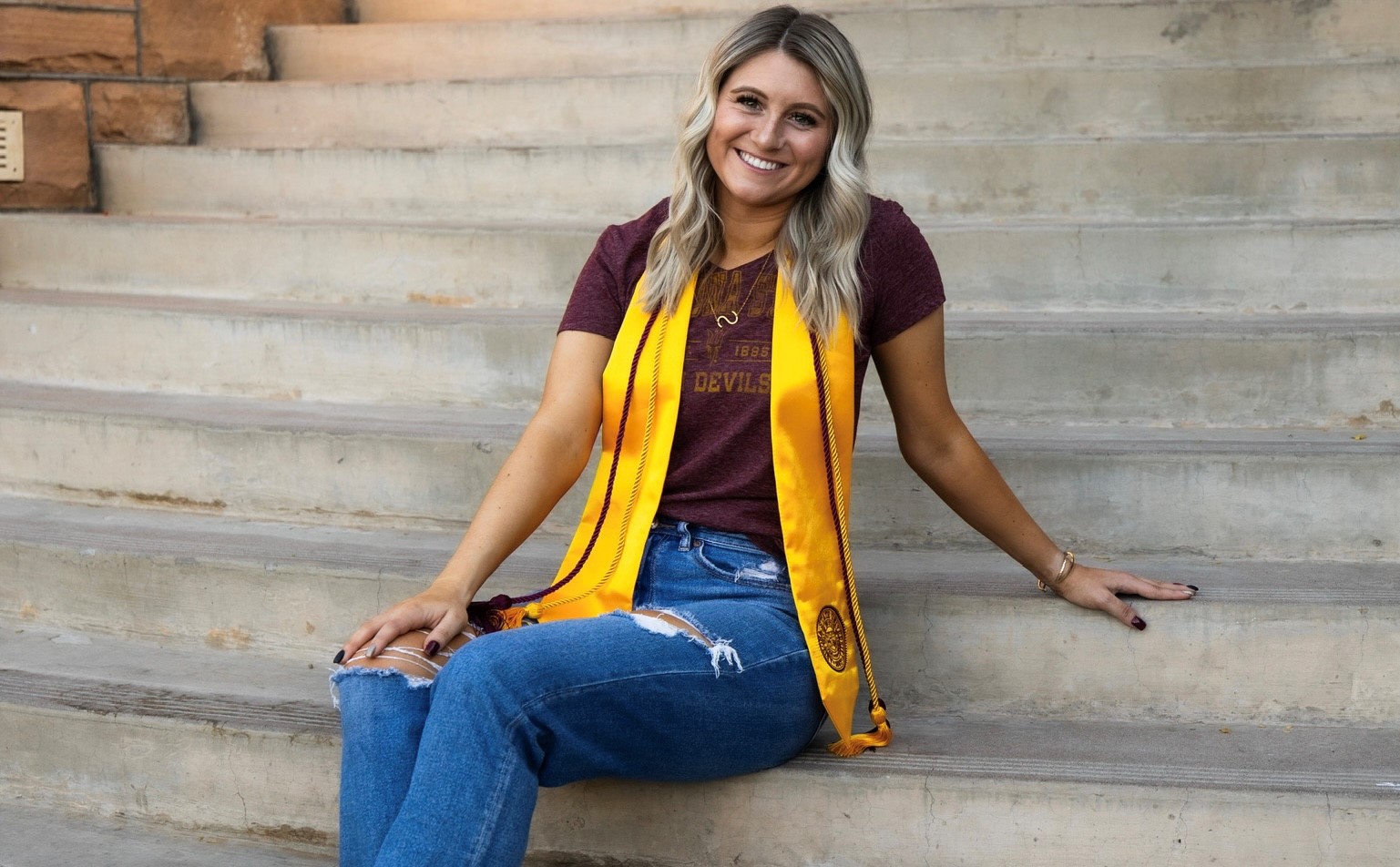  sits on concrete steps, smiling at the camera with her gold graduation sash and maroon cords draped around her neck