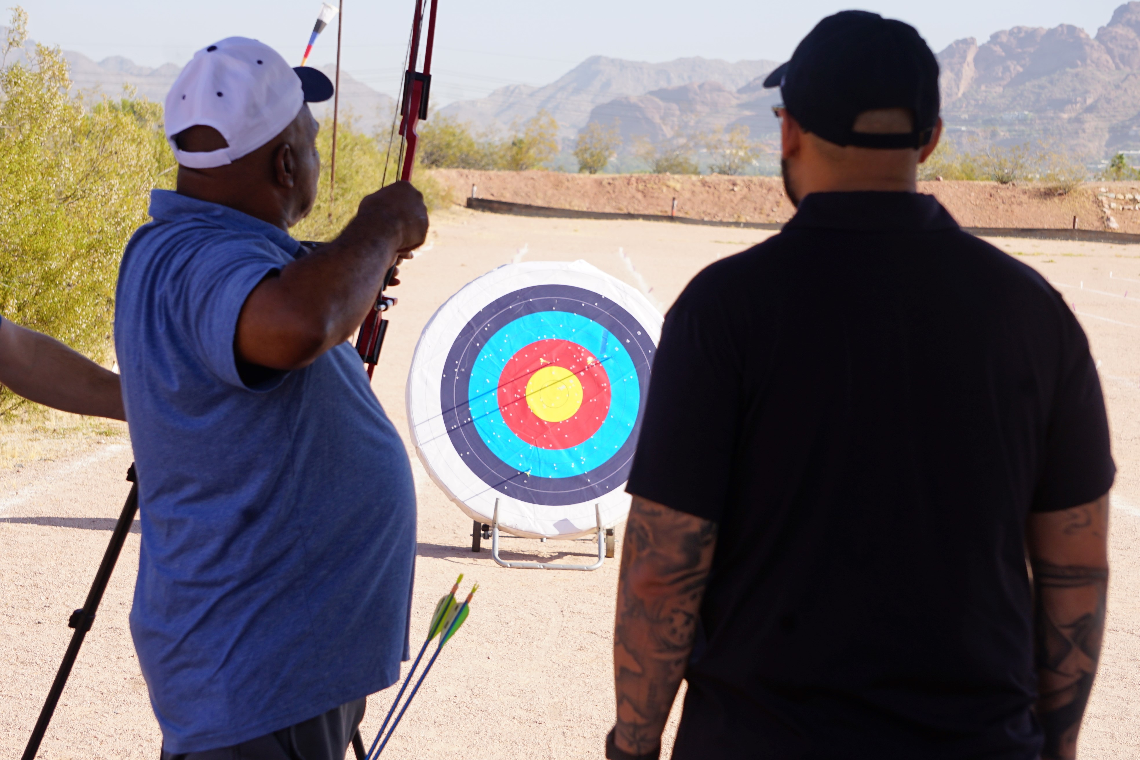 James Malone stands behind a target holding a bow and arrow as Rick Alvarado watches