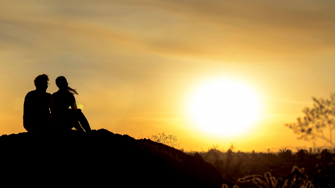 A silhouetted man and women sit on a mountain top looking out at a golden sunset.