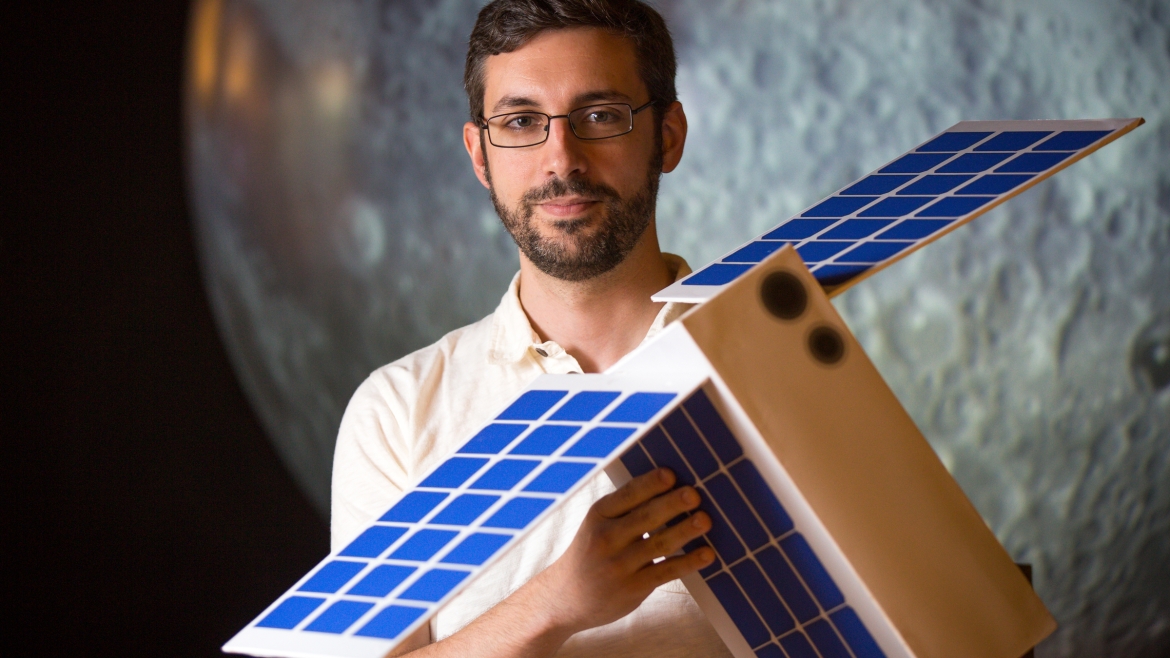A man holds a model of a shoebox-size satellite.