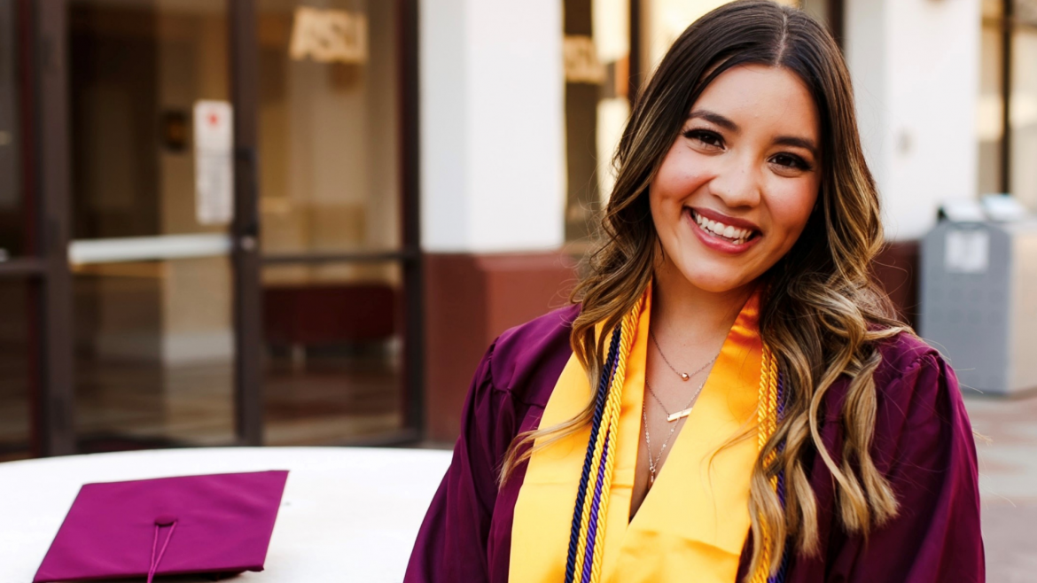 Ana 'Juli' Rincon smiles at the camera. She's wearing her maroon graduation gown and gold graduation stole and cords. Her cap is on the table next to her