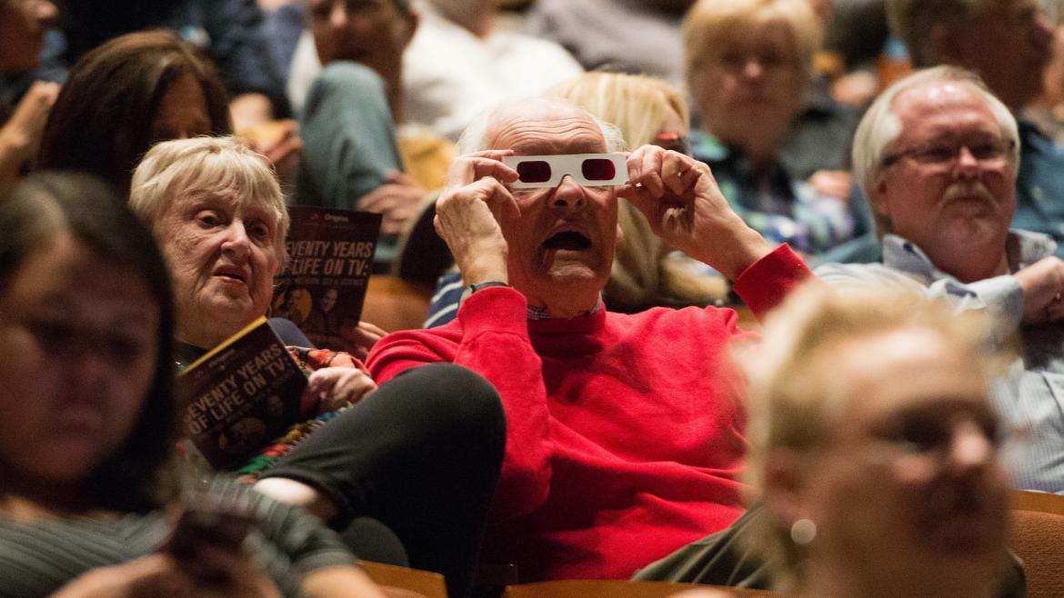 A man in a theater audience wears 3-D glasses.