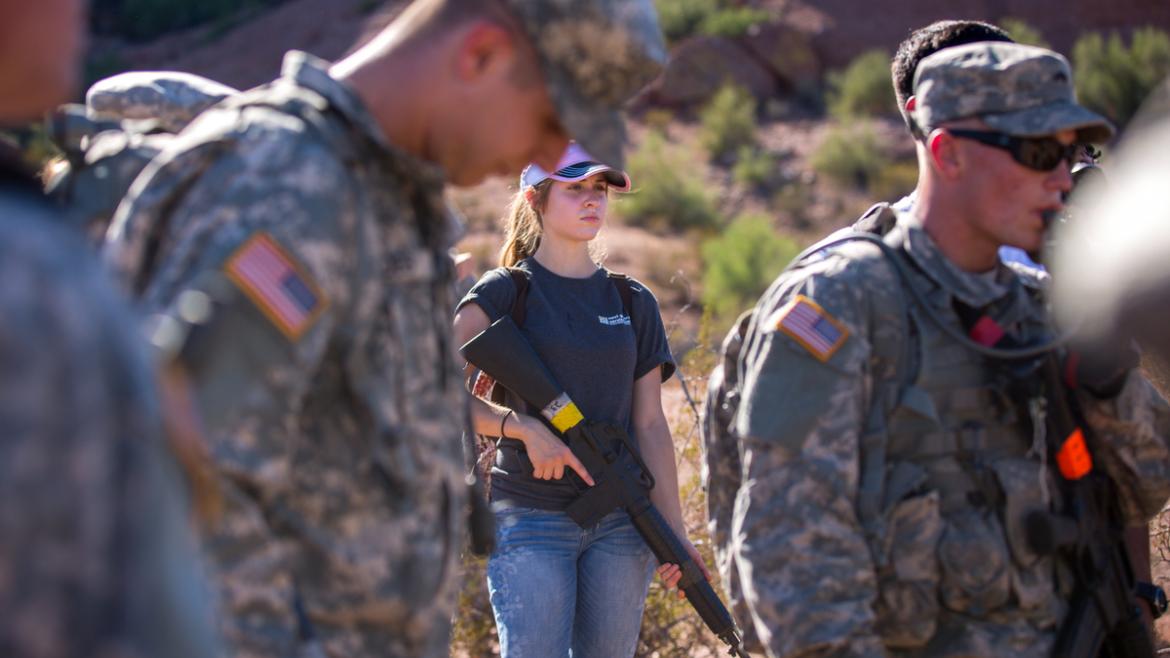Woman holds a dummy riffle near military men.