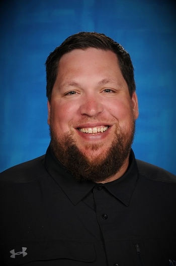 Portrait of ASU alum Colin Donovan. He has short black hair and a beard and smiles at the camera in front of a blue backdrop.hirt