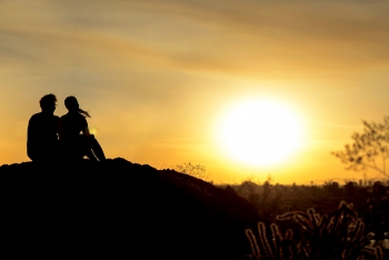 A silhouetted man and women sit on a mountain top looking out at a golden sunset.