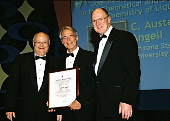 Three older adult men standing on a stage wearing tuxedos and bow ties. The man in the middle holds a framed award.