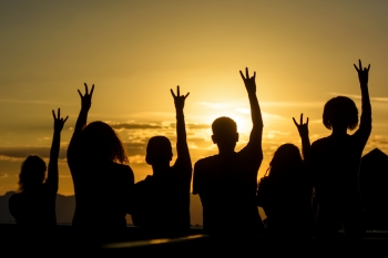 Group of people making ASU Pitchfork signs with their hands, silhouetted against a background of a setting sun.