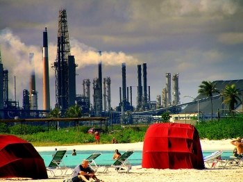 People sitting on a beach in the foreground while industrial smokestacks loom in the background.