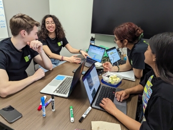 Students gathered around a table with laptops mid-discussion.