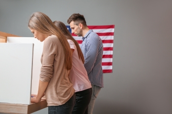 Young adults voting at booths next to a wall with an American flag.