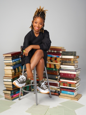 Alena Wicker sitting on a chair with stacks of books behind her.