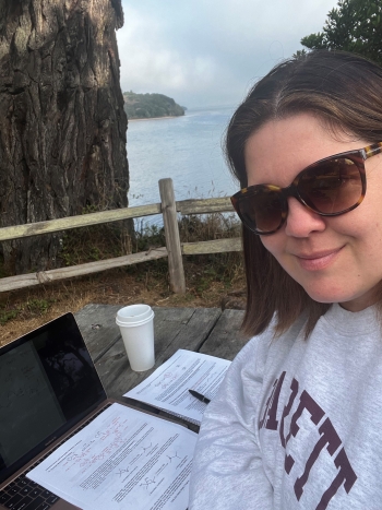 Portrait of ASU Online student Shawna Brechbill wearing sunglasses and sitting by the ocean with her laptop, some paper and a coffee.