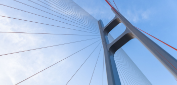 A gray suspension bridge is viewed from below with a blue sky behind it.