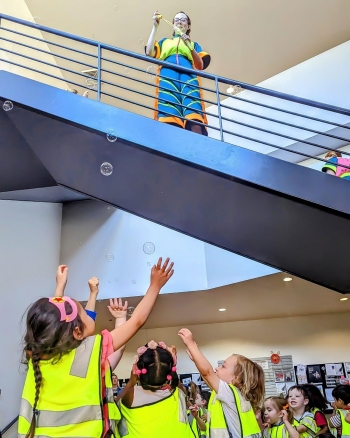 Person stands on staircase blowing bubbles and children below reach up to catch them.