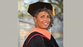 Dawn Augusta smiles for the camera. She wears her doctoral regalia with her black cap, gold tassels, and black gown with maroon trimmings. 