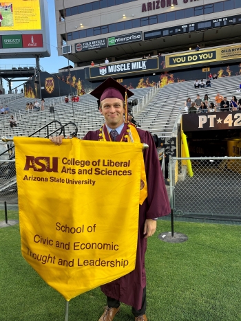 Joe Pitts in his graduation robe and cap holding a sign for the School of Civic and Economic Thought and Leadership