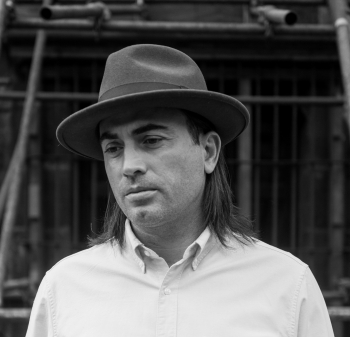 Black and white photo of A.S. Dillingham who has shoulder-length dark hair, a short-brimmed hat, and is wearing a white button-down shirt.