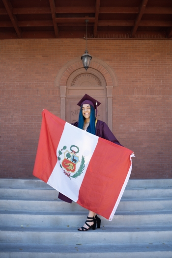 ASU alum Stefania Becerra Lavado wearing a maroon cap and gown and holding the Peru flag on the steps of ASU's Old Main building.