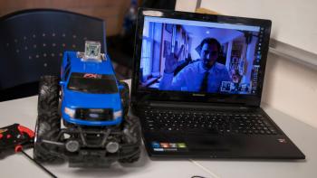 A student poses on a laptop webcam feed next to a remote controlled car during a project demonstration.