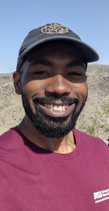 Close up image of PhD graduate Ed Buie II wearing a ball cap, smiles in front of a desert landscape.