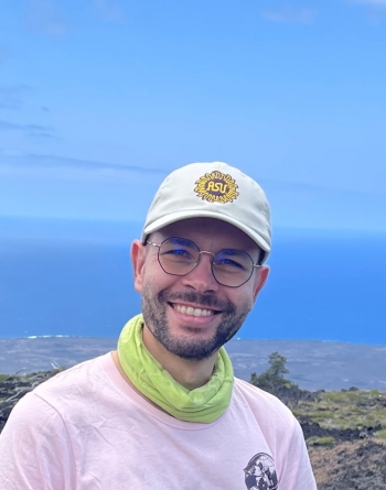 Florian smiling at the camera wearing an ASU hat, a pink shirt and a green neck gaiter with an ocean in the background